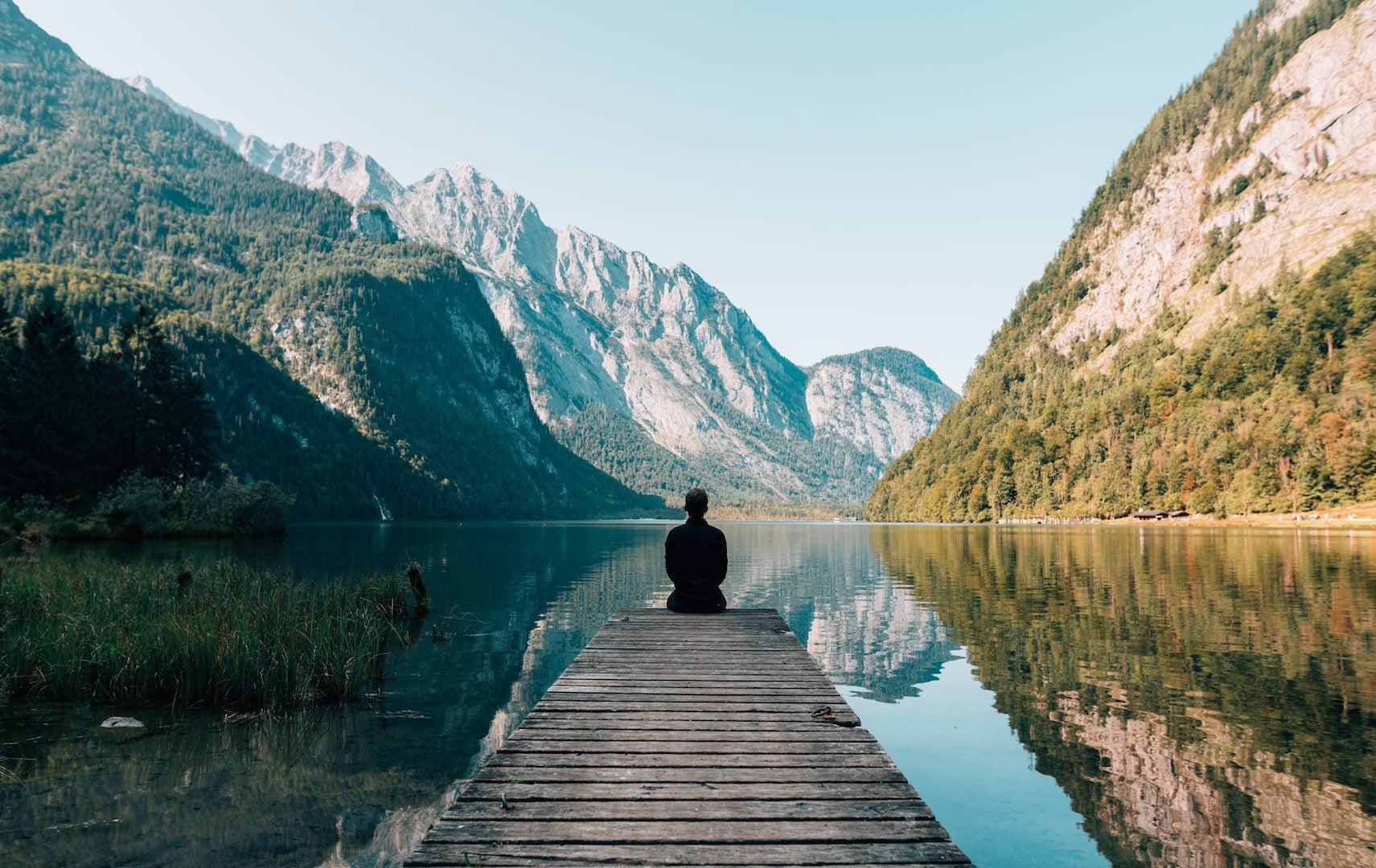 A man sitting on a dock near a lake surrounded by mountains.