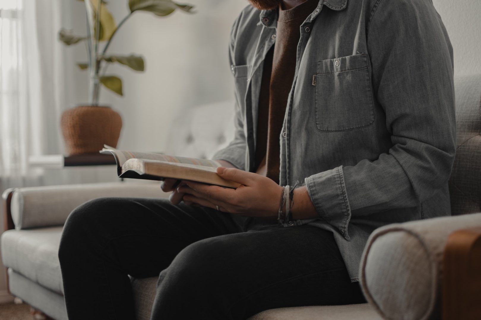 A man sits on the couch while reading the Bible.