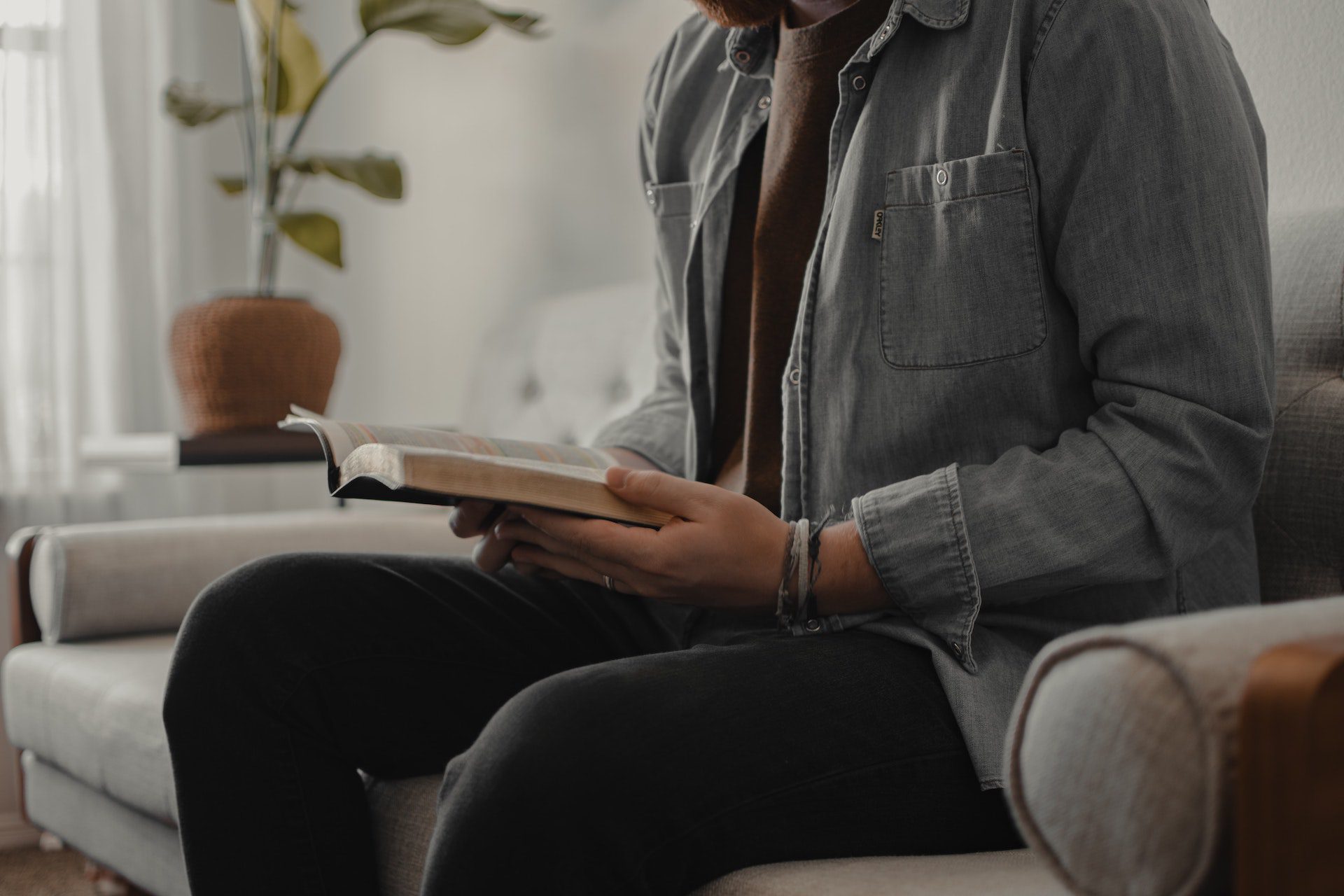 A man sits on the couch while reading the Bible.