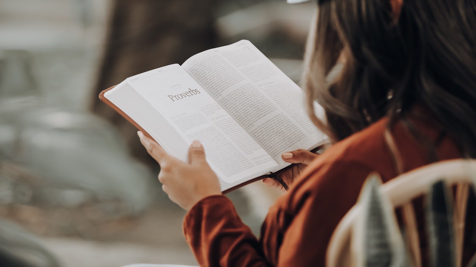 A woman with her Bible open to Proverbs, while sitting on a chair outside.