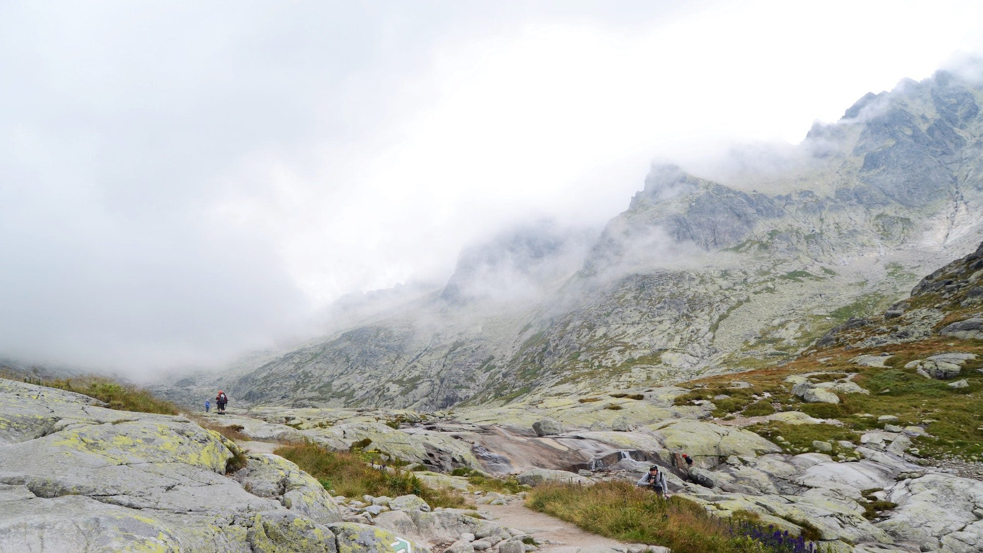 People walking on rocks at the base of mountains on a cloudy day.