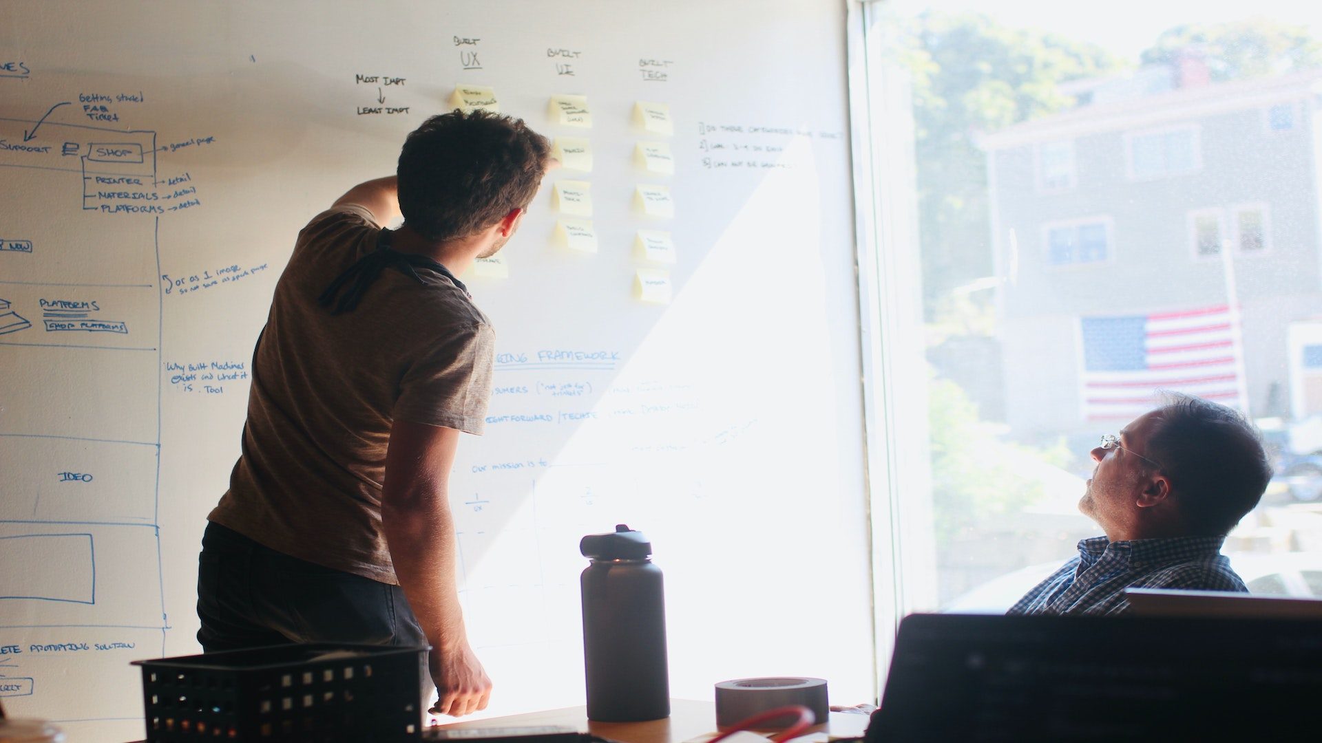 Two men in a business meeting, with one man sitting at a table in an office and the other man writing on a whiteboard.