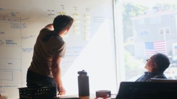 Two men in a business meeting, with one man sitting at a table in an office and the other man writing on a whiteboard.