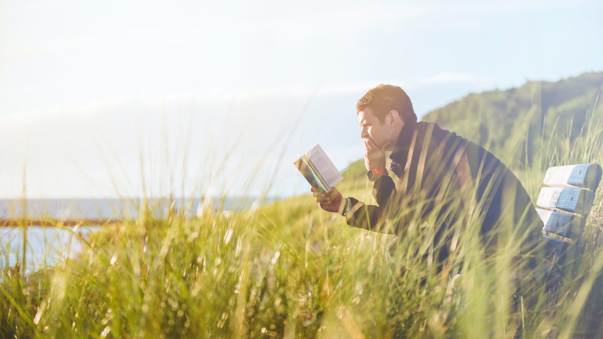 Man reading the Bible on a bench by the beach.