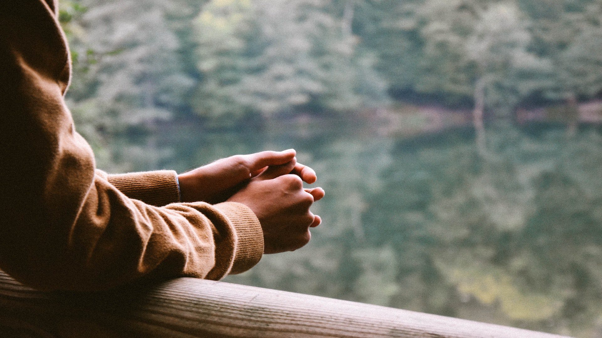 A person's hands folded over a railing near a lake.
