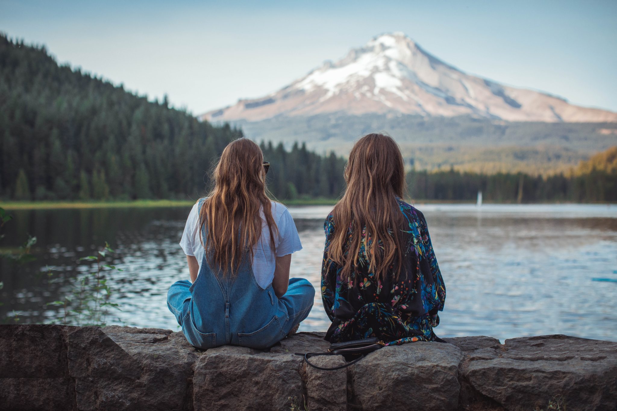 Two women sitting on a rock wall overlooking a lake.