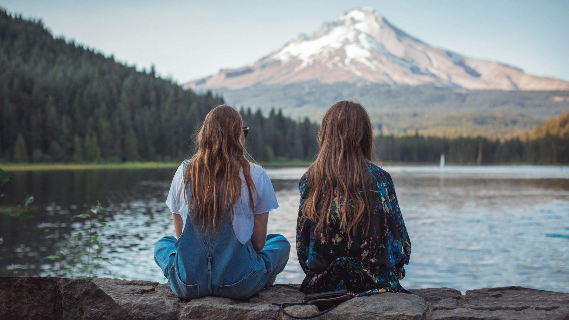 Two women sitting on a rock wall overlooking a lake.