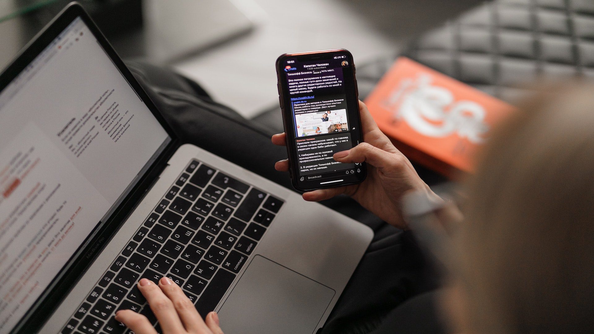 Young woman on leather couch working at a laptop while browsing her phone.