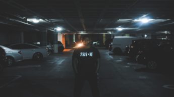 A man standing in a parking garage with a shirt that says Jesus is greater than me.