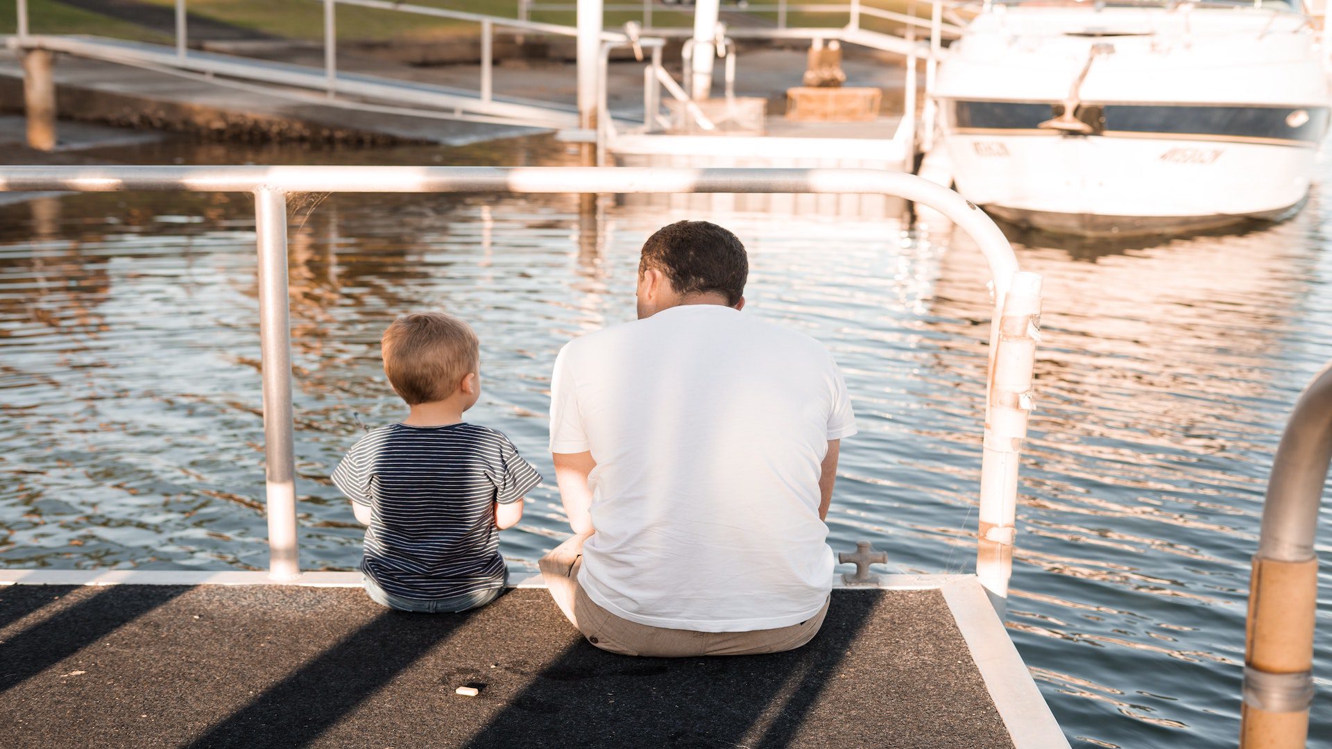 Dad sitting with toddler boy on a dock near water and boats.