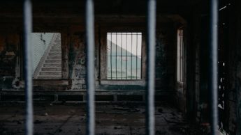 Prison bars in front of an empty room with a window with bars.