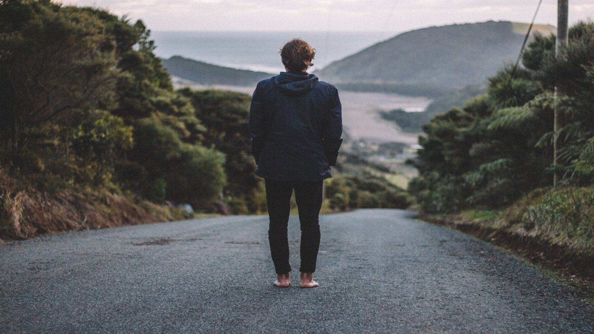Man standing barefoot on a road overlooking mountains and water.