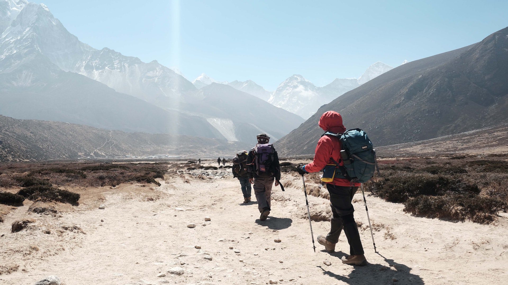 A group of people hiking with backpacks on a dirt bath at the base of mountains.