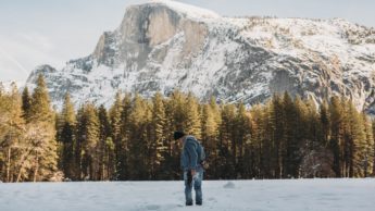 Man in jeans and denim jacket standing on snowy field in front of a forest and mountain.