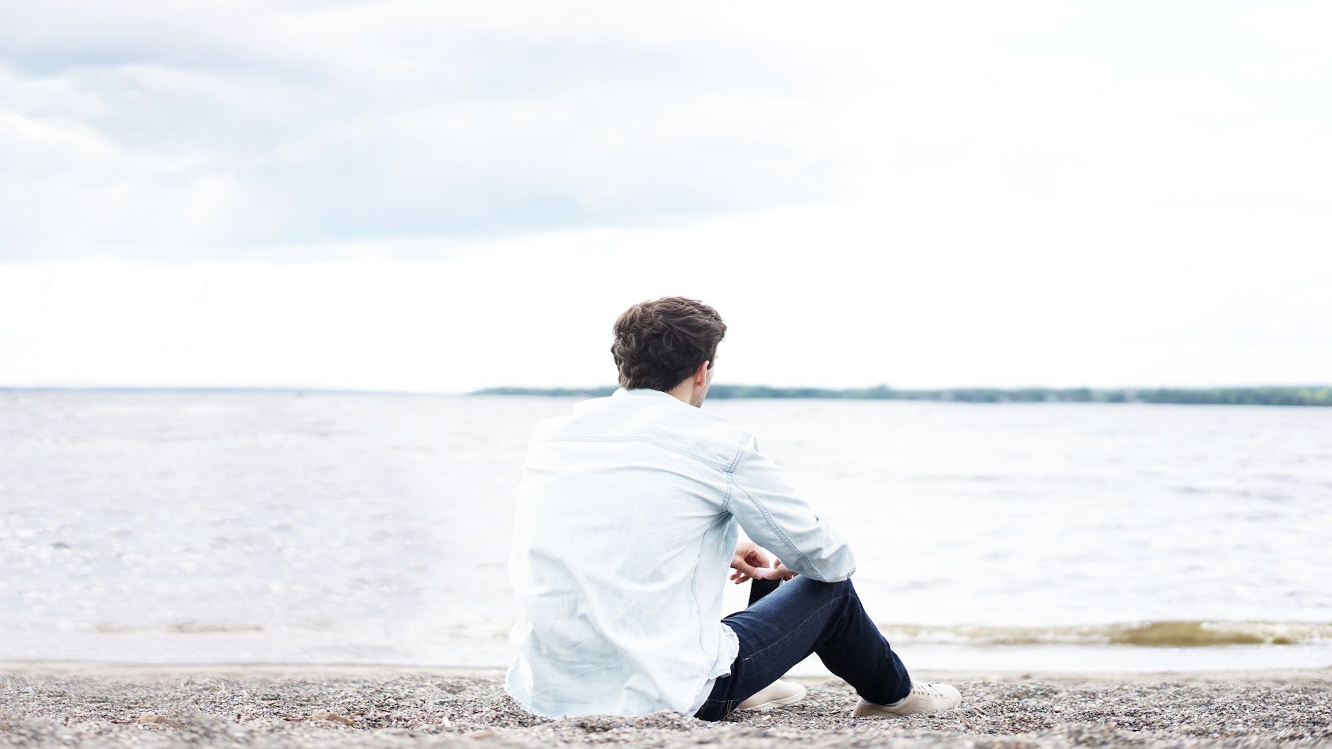 A man in jeans and white shirt sitting on sand in front of a lake.