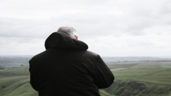 Back of an elderly man in a black hooded jacket standing over a green valley.