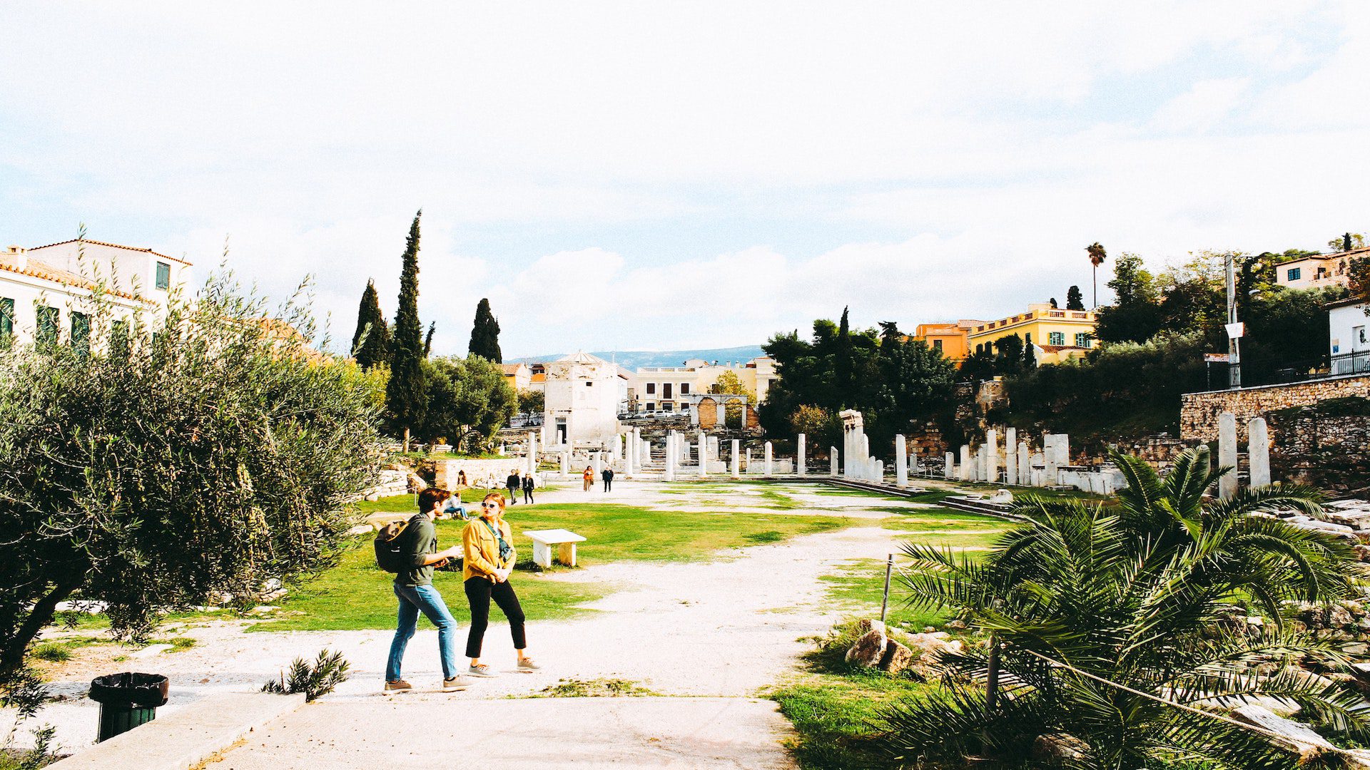 A man and a woman walking outside on a dirt path near houses and trees.