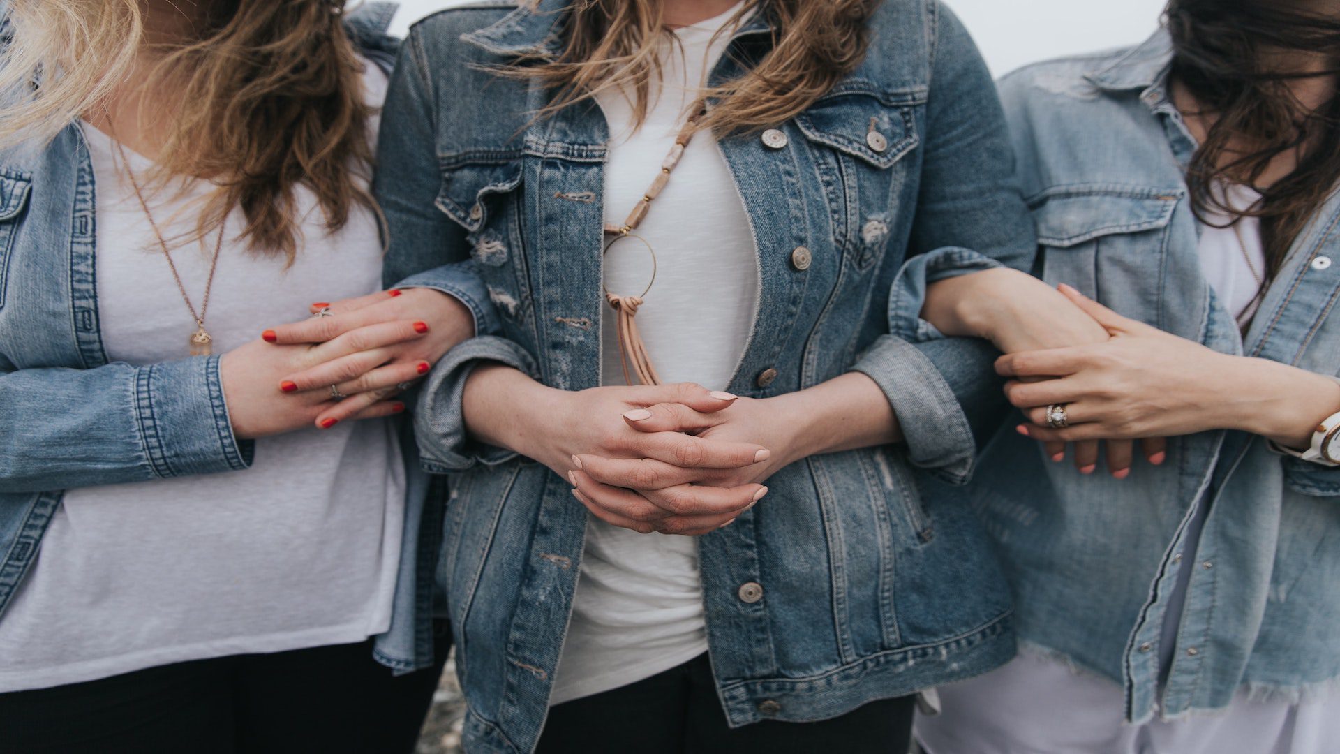 Three women in white shirts and denim jackets with their arms linked together.