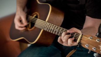 A man in jeans and a black t-shirt sitting on a coach while playing an acoustic guitar.