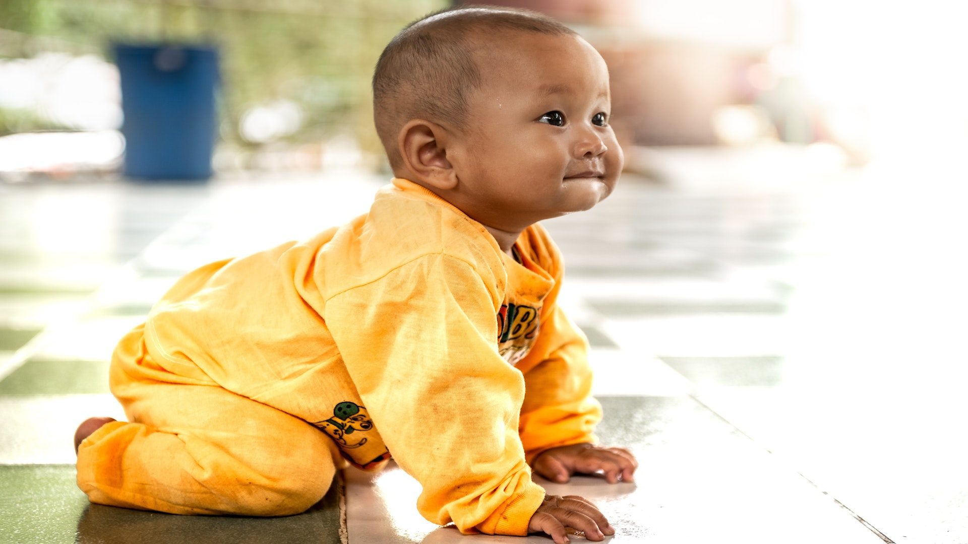 Baby boy in a yellow onesie crawling on the floor.