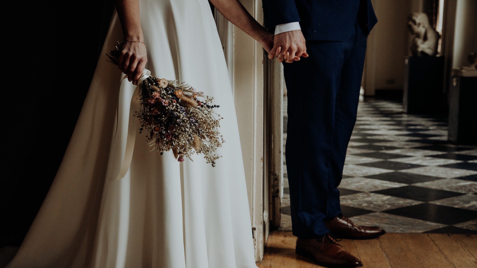 A man in a suit and a woman in a wedding dress holding hands around a corner.