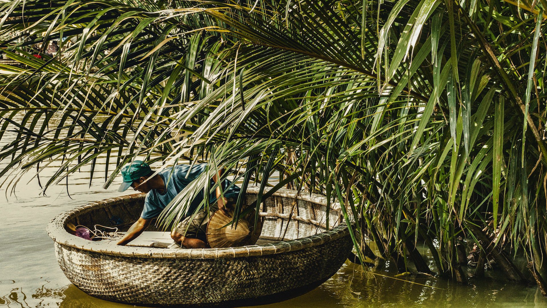 A man in a boat under green leaves on a river.