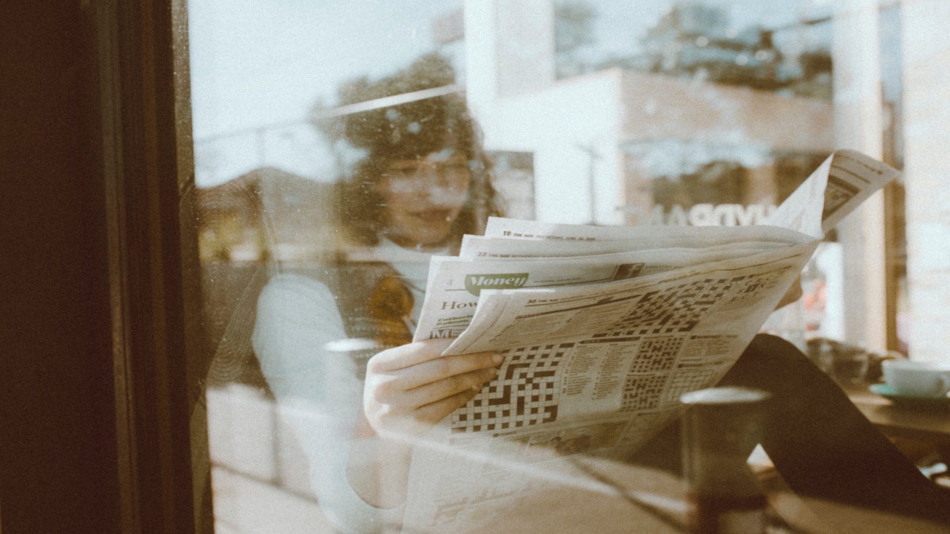 A woman reading a newspaper at a cafe through a window.