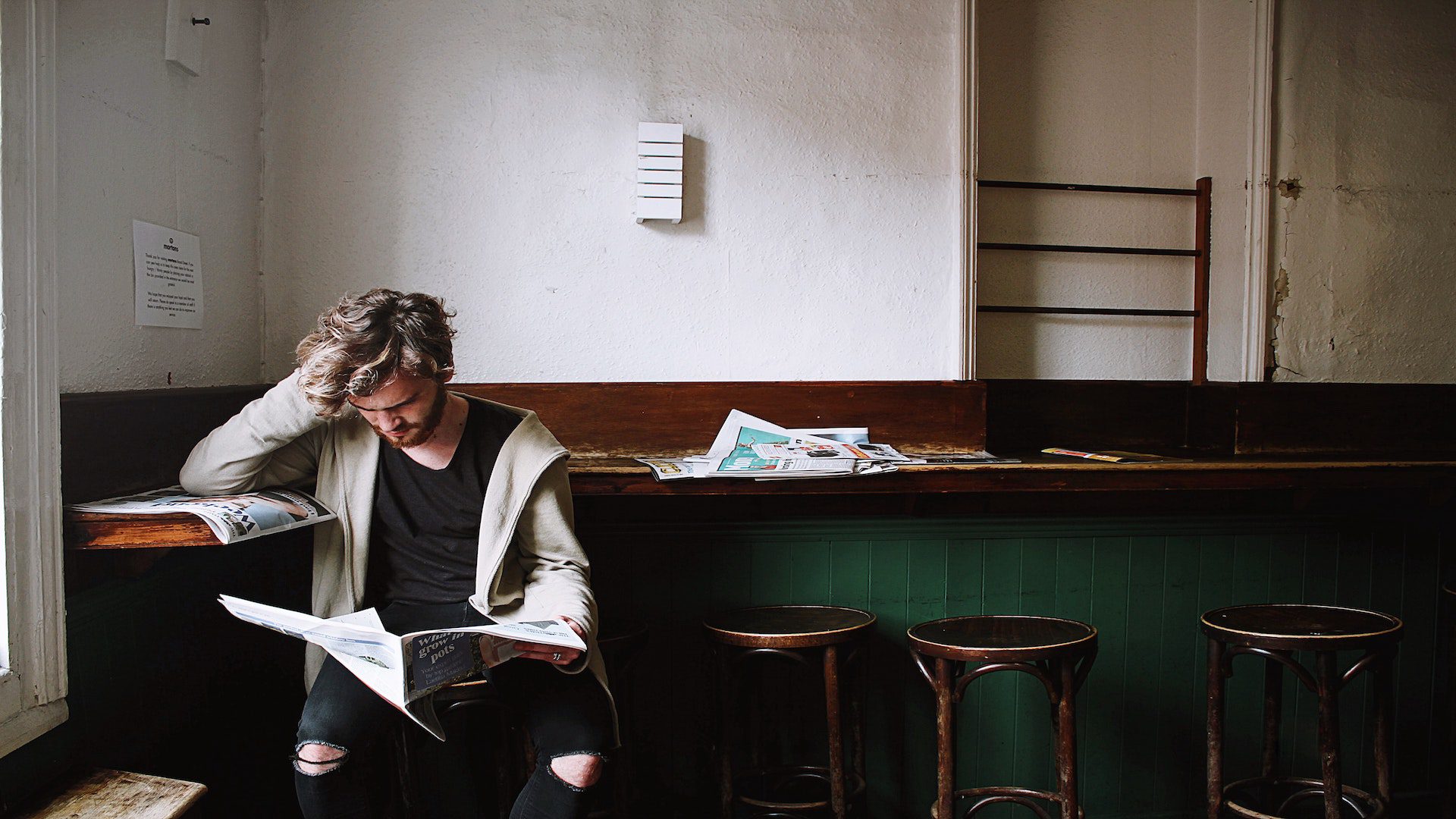 A man sitting on a bar stool in a cafe reading a newspaper.