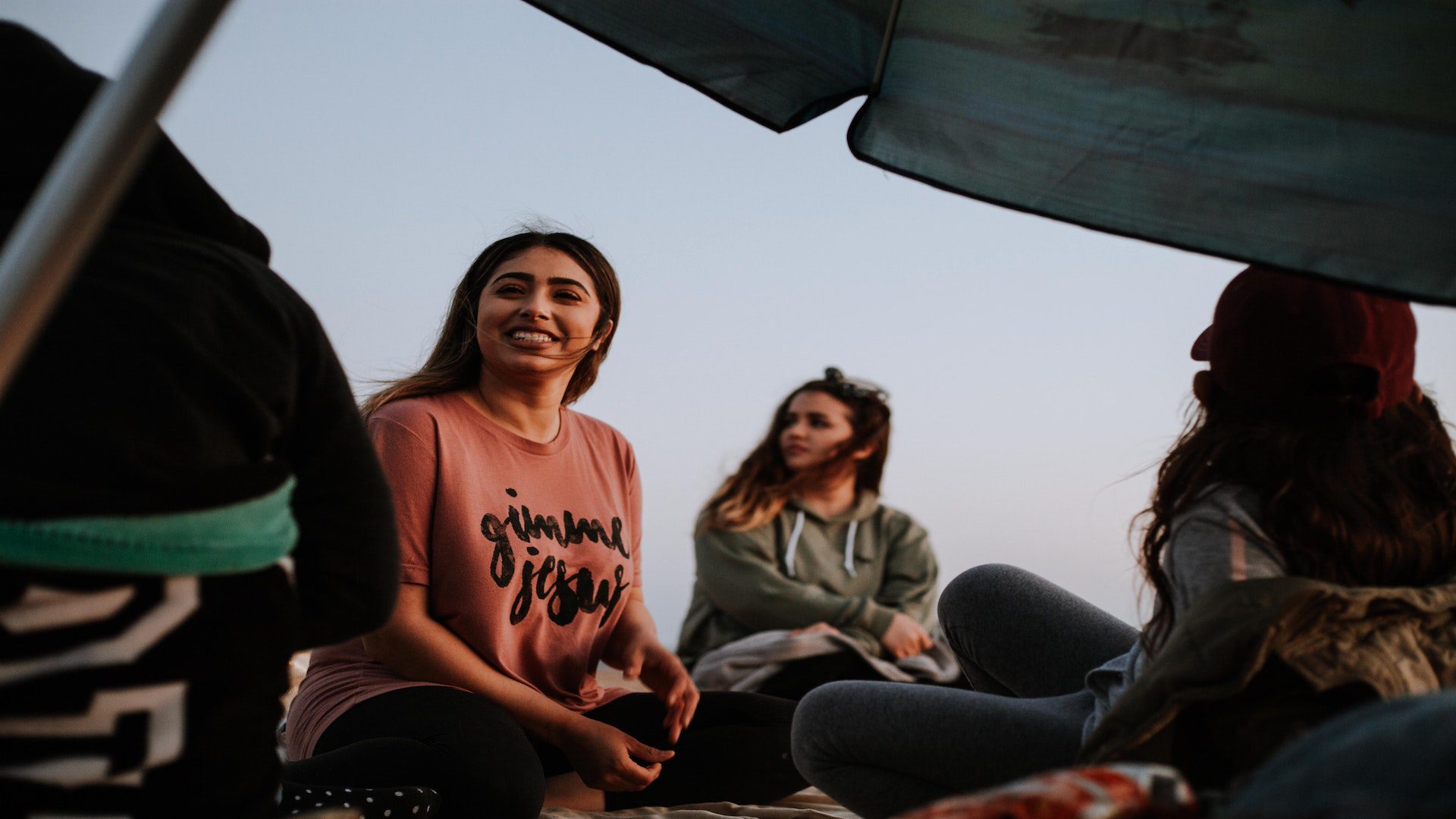 Three woman sitting on the ground outside under an umbrella.