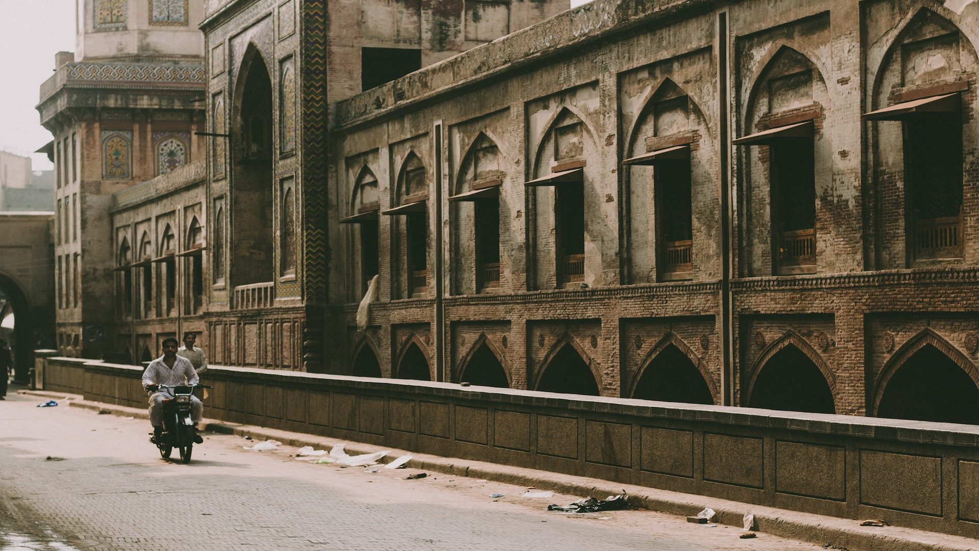Man riding a motorcycle with a man walking behind him on an empty street next to a brick building in Pakistan.