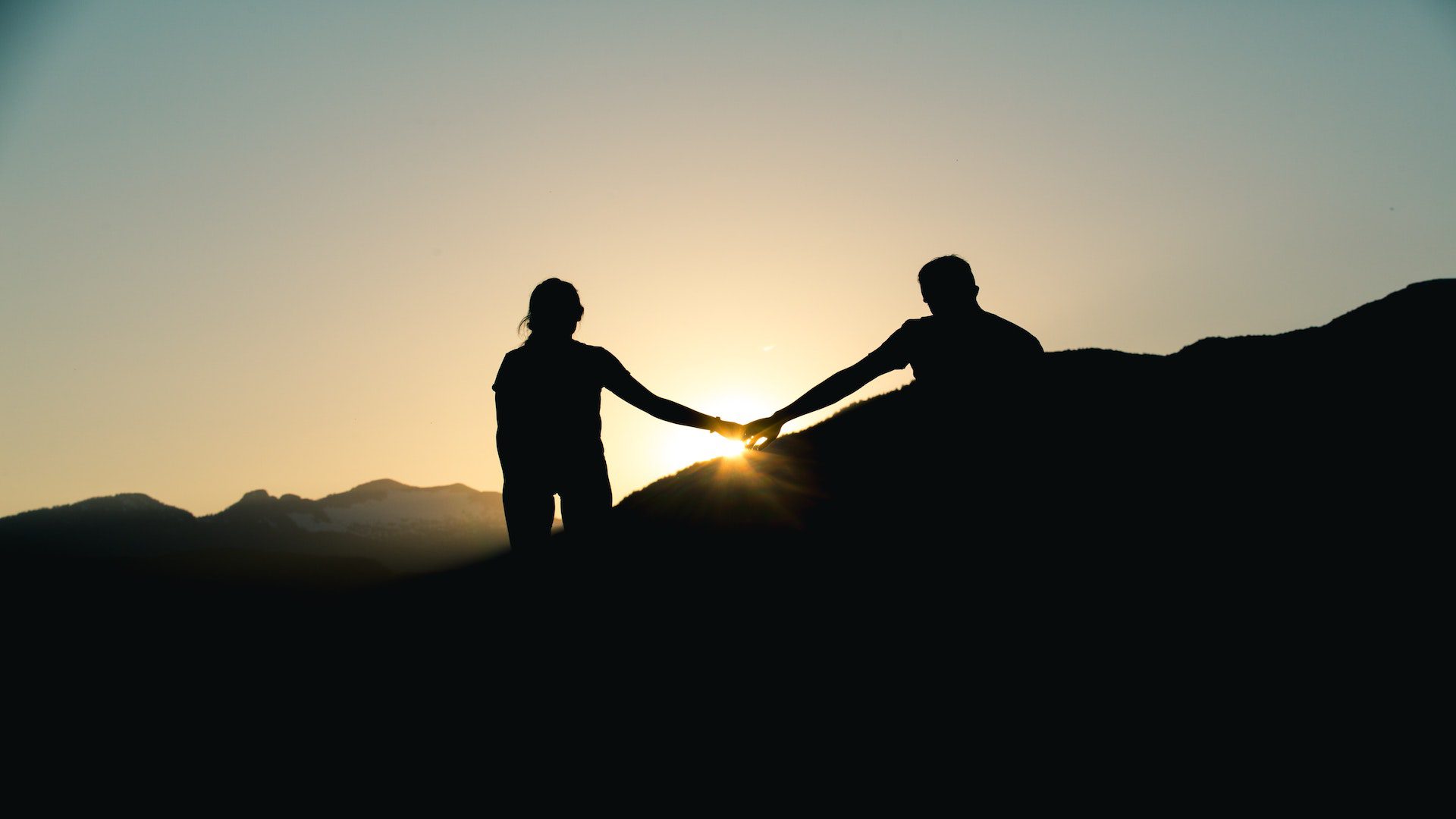 Silhouette of a woman and man holding hands on a mountain during a sunset.