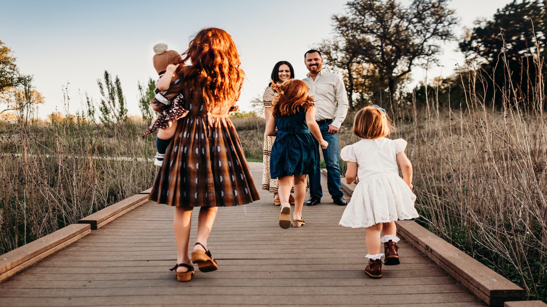 Three young girls, one holding a baby, running towards their parents on a sidewalk.