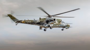 Military helicopter with gray and green camo flying in the air.