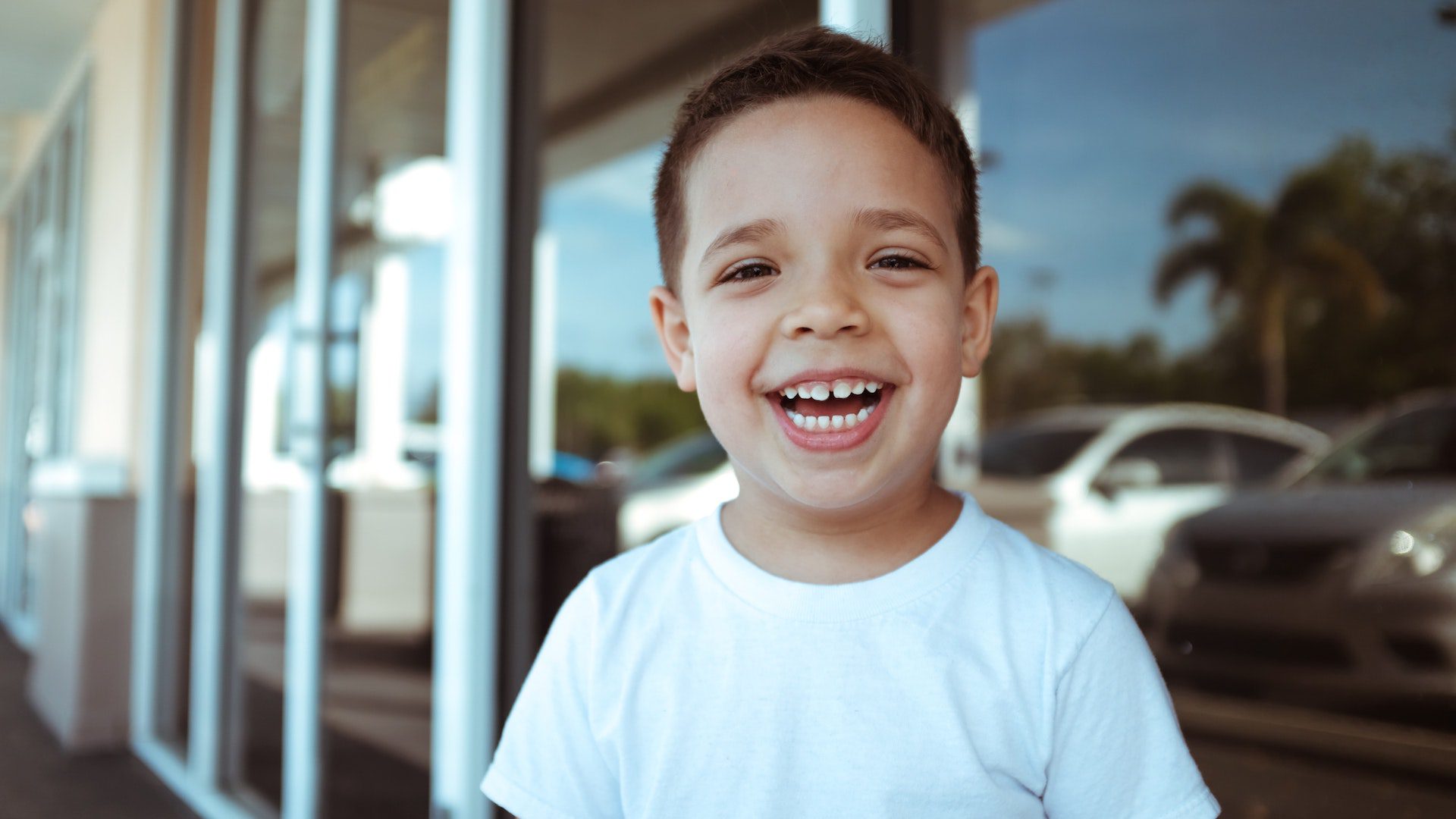 Young boy in a white t-shirt smiling outside in front of the windows of a store.