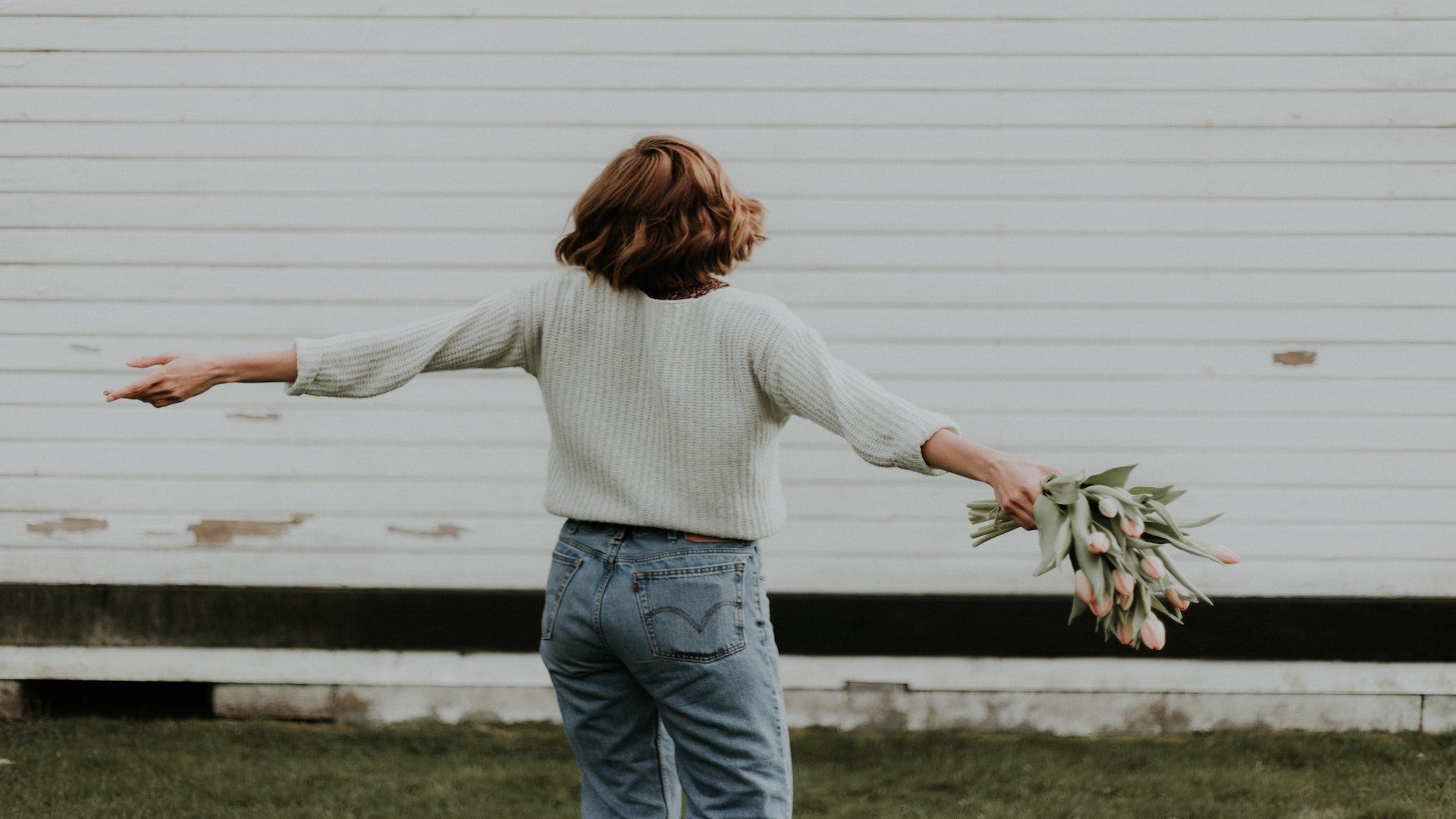 Back of a woman in a white sweater and jeans holding flowers standing in front of a white house.