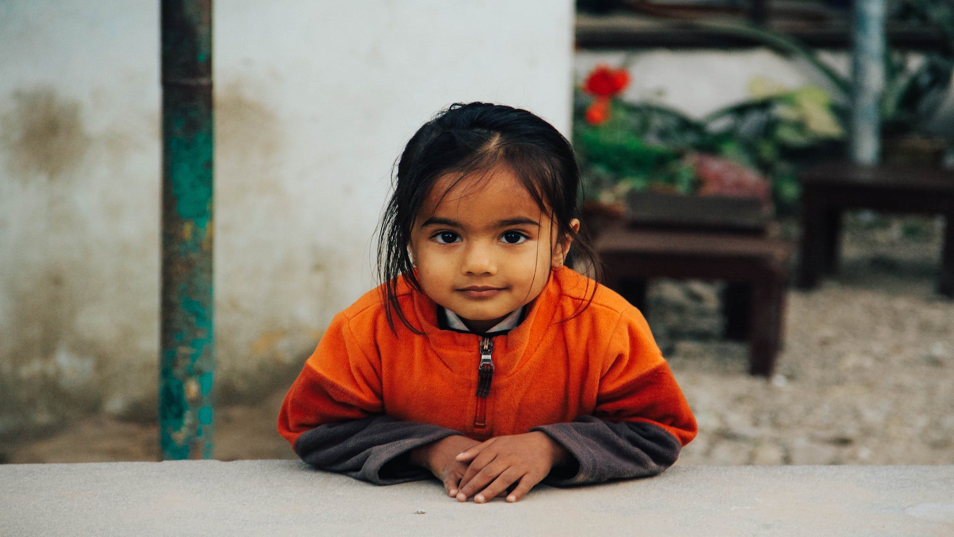 Young girl with dark hair and an orange sweatshirt leaning over a table outside.