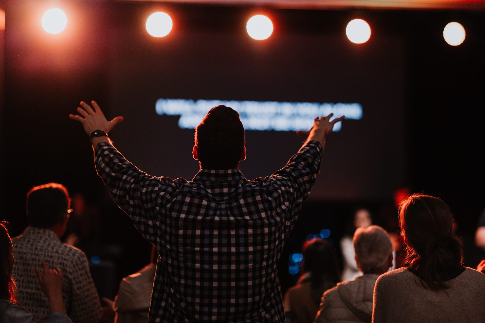 A man with his back turned towards the camera raises both of his arms as he sings in worship