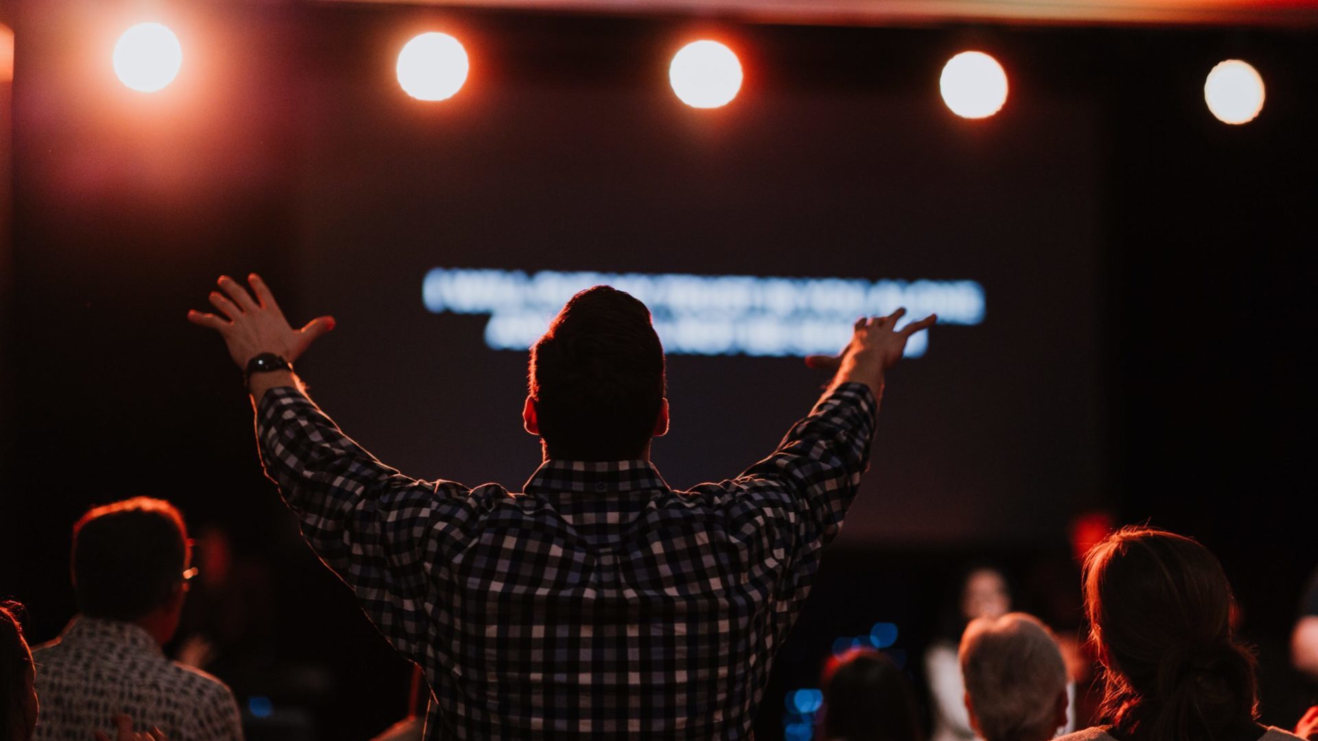 A man with his back turned towards the camera raises both of his arms as he sings in worship