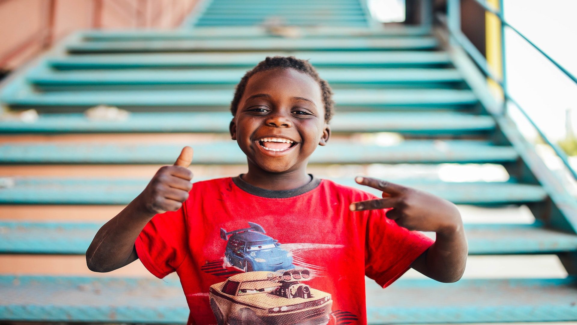 Young boy in Africa standing outside in front of stairs in a red t shirt holding a thumbs up and peace sign.