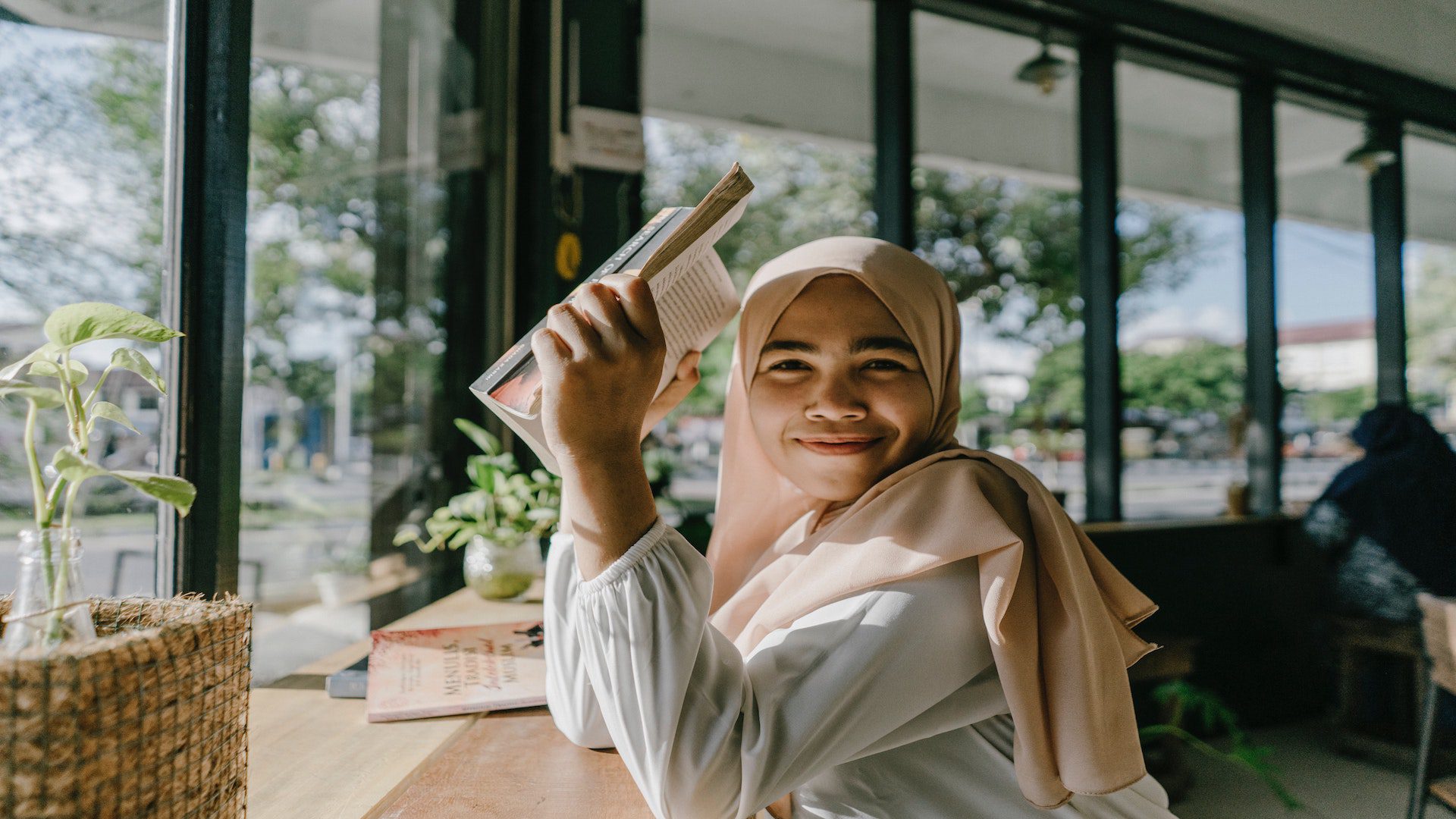 Woman in a pink hijab holding a book and sitting at a table in a cafe in front of a window.