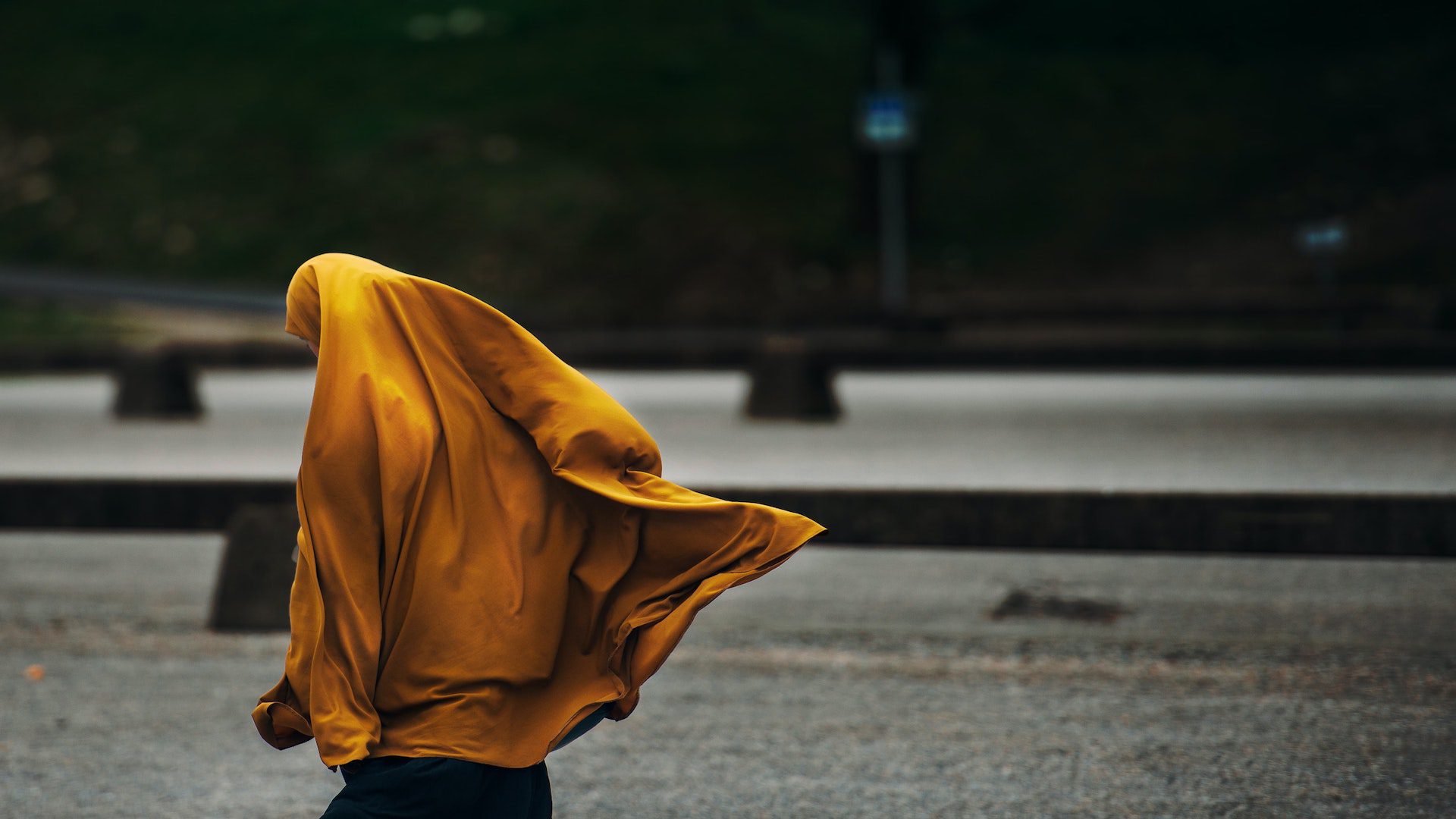 The back of woman with a yellow hijab and black skirt walking on a street.