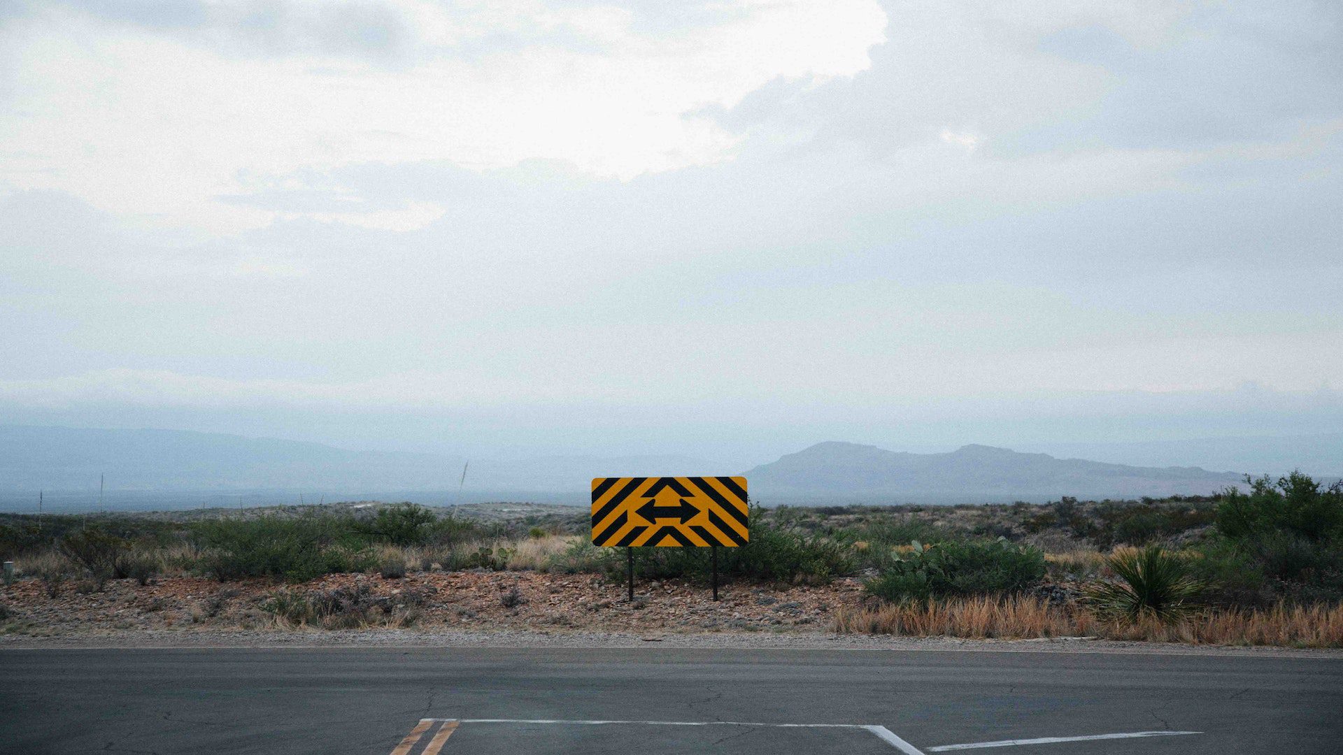 A black and yellow sign with an arrow pointing both ways at the end of a road. There are mountains in the background on a cloudy day.