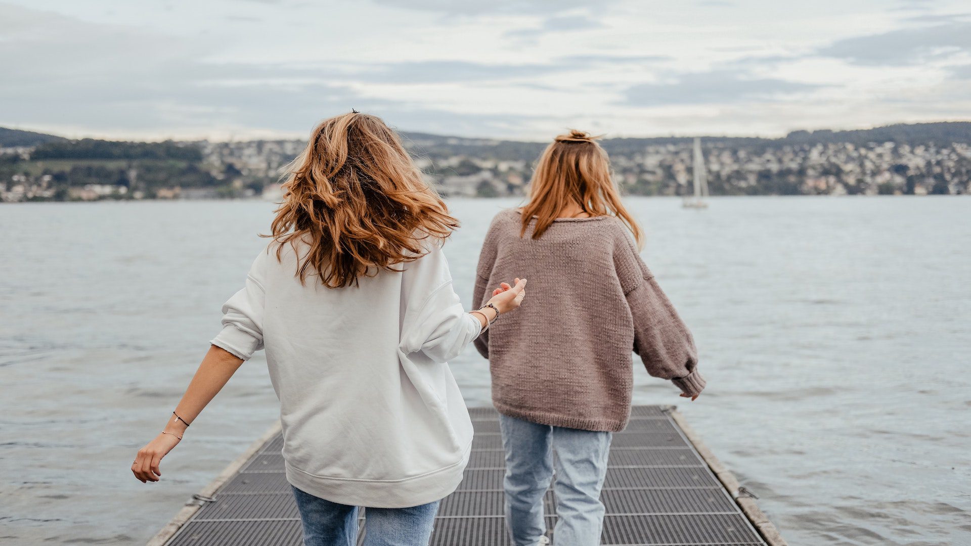 The backs of two women walking on a dock near a lake on a cloudy day.