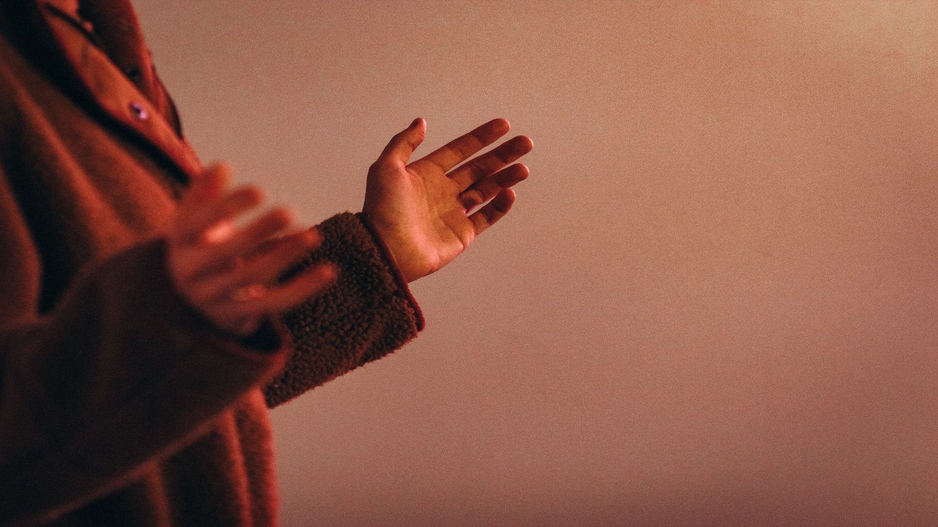 The side of a woman in a red coat with her arms raised in front of a pink wall.
