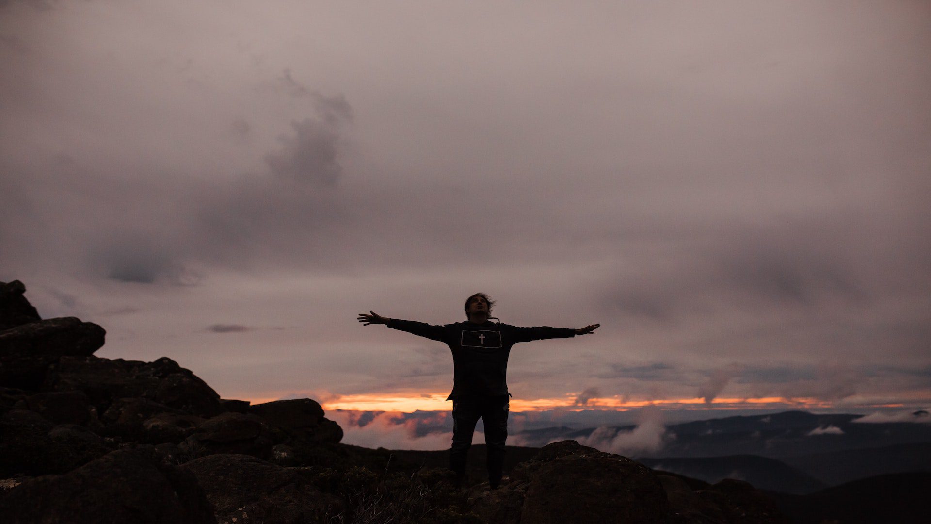 A man with black pants and a black sweatshirt with his arms raised standing on rock during a sunset.