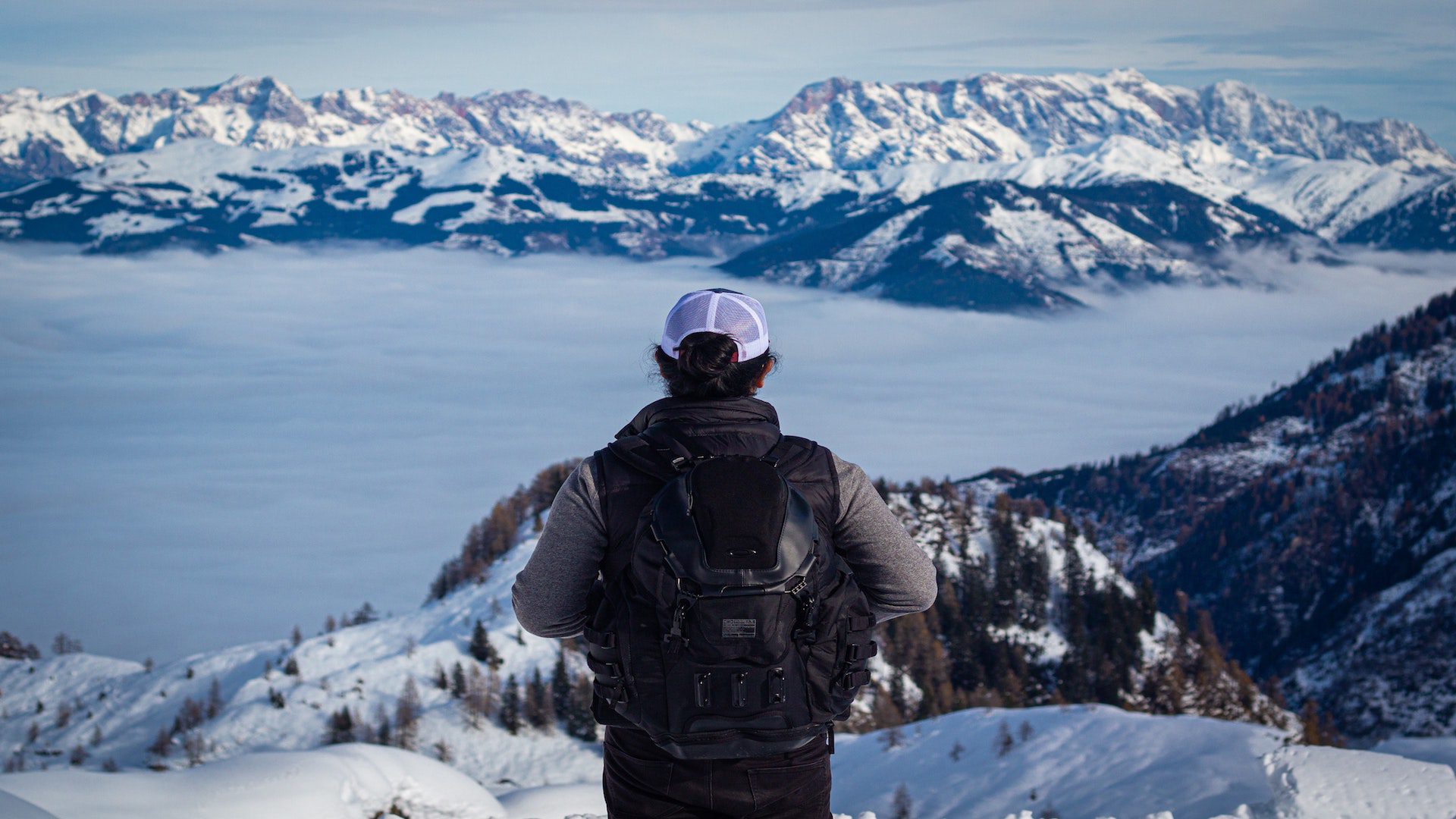 The back of a person in a black vest with a back backpack standing on the edge of a snowy mountain.