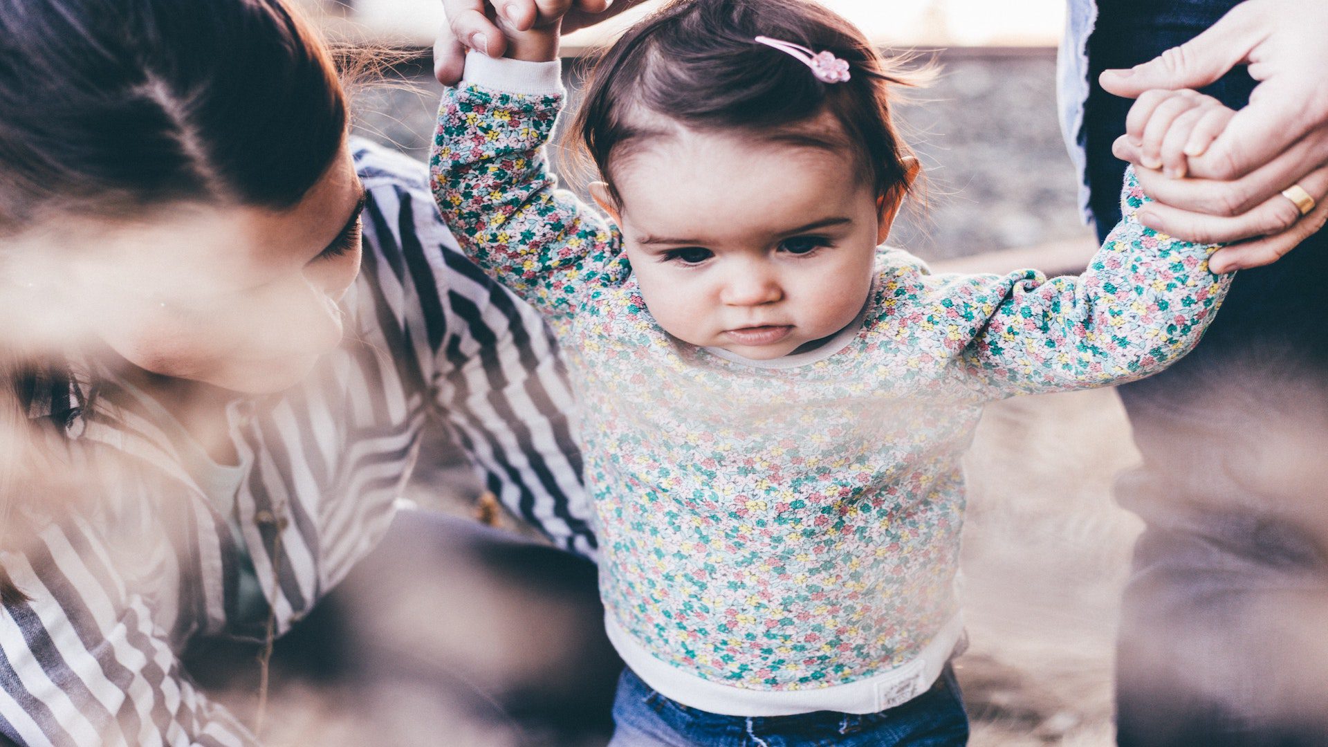 A baby girl in a floral shirt walking and holding the hands of a person behind her. There is a woman kneeling besides the baby.