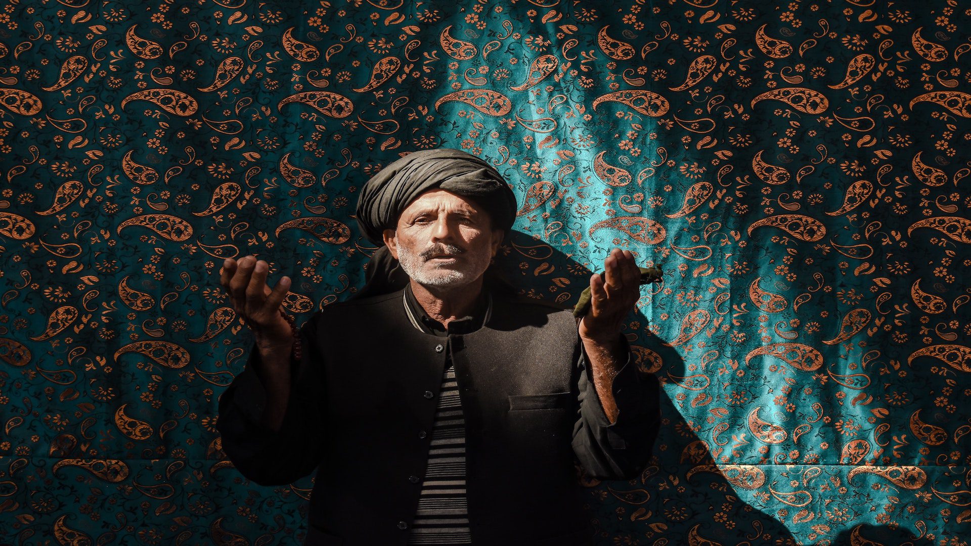 An elderly Iranian man in a black coat and a turban with his arms raised. He is standing in front of a blue and gold patterned tapestry.