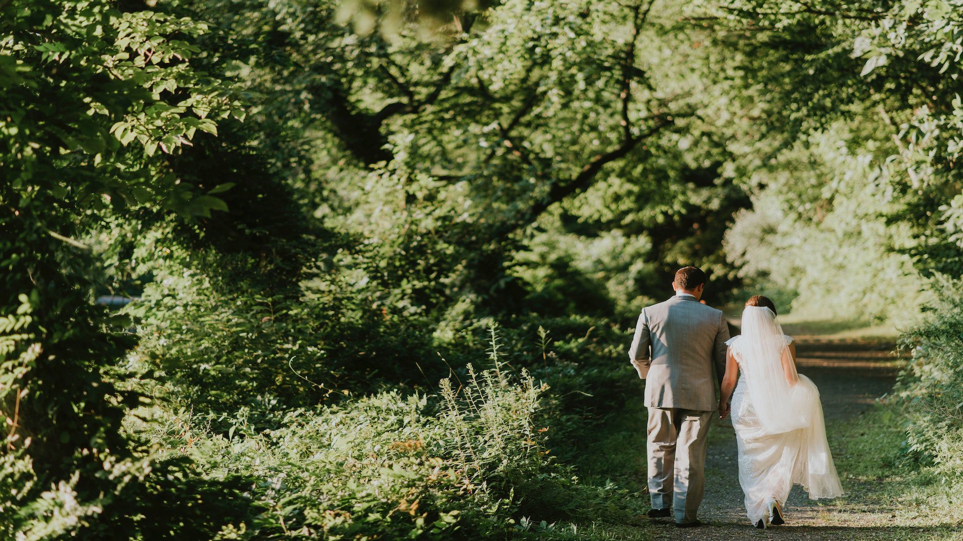 A bride and groom walking on a grass path surrounded by green trees.