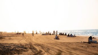 People sitting and standing on sand at a beach on a sunny day.